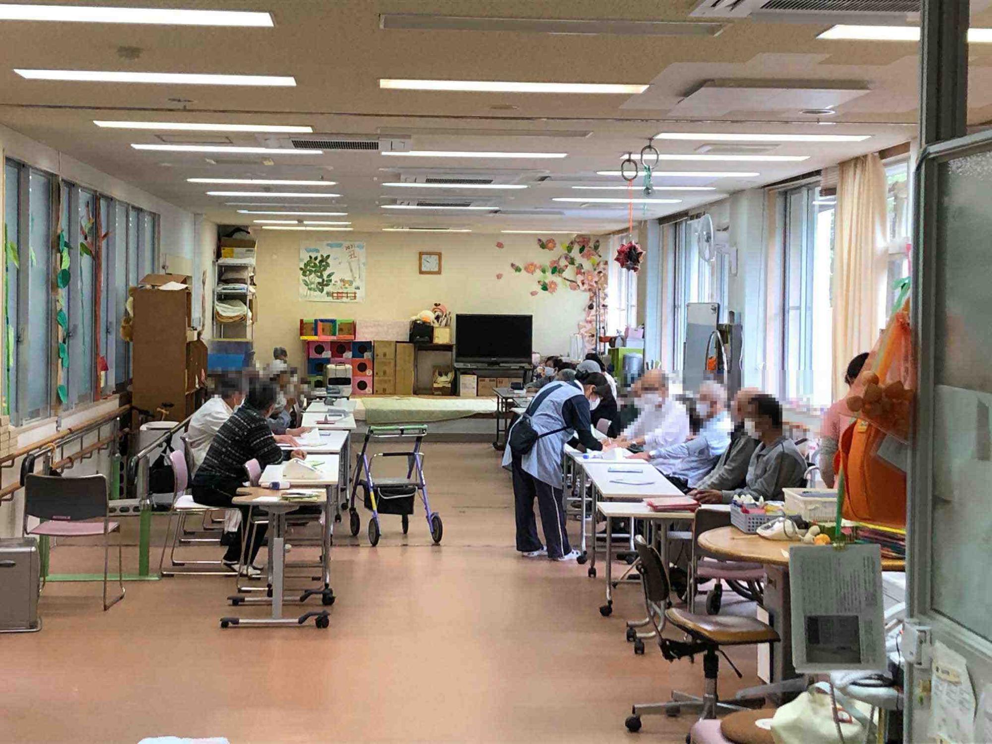 向い合うように設置された机の席につきデイケアに参加している高齢者の方々の写真