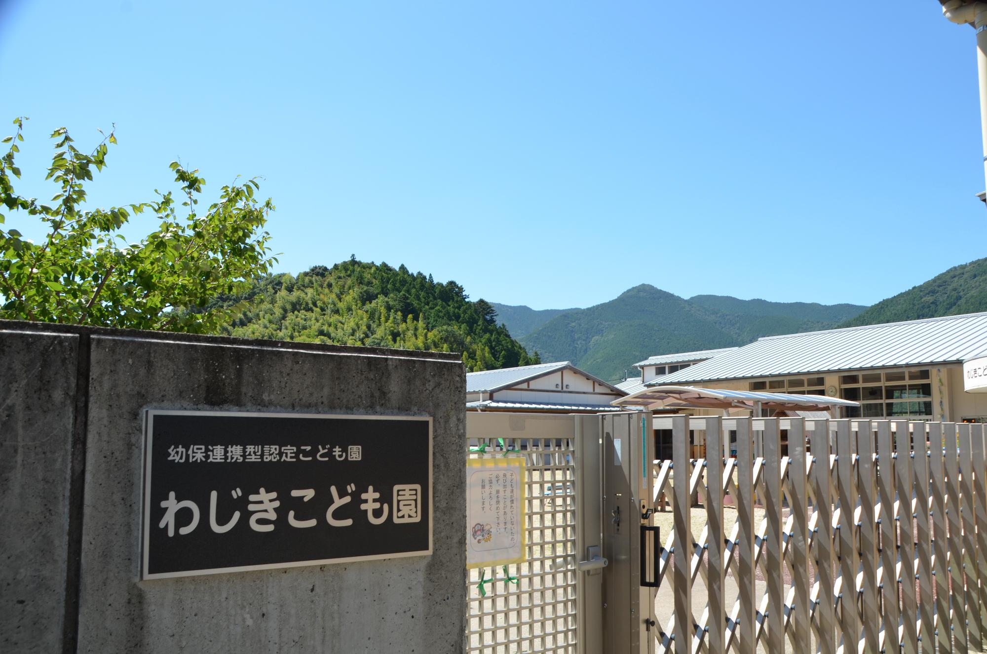 雲一つない青空の下、「わじきこども園」と書かれた園銘板が設置された外壁の写真