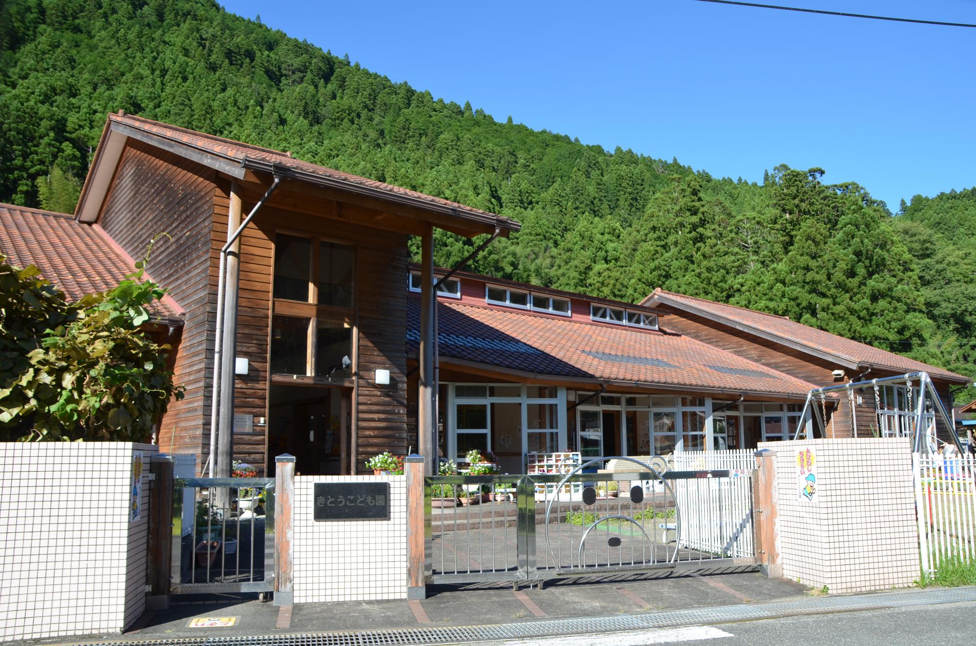 雲一つない青空と青々とした木々が生い茂っている山を背景に、「きとうこども園」と書かれた園銘版と茶色い屋根の木製のログハウスのような園舎の写真