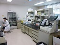 薬剤の入ったガラス窓の棚、中央の机には機材や備品が並んだ棚が置かれ、奥の机で女性の職員が作業をしている検査室の様子の写真