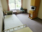 奥に外の景色が見える窓があり、左側にベット、右側にテレビの置かれた棚、窓際に机と椅子が設置されている入院個室の写真