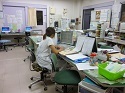 パソコンが設置された机が奥にあり、中央に並んでいる机では、看護師の女性がパソコンの前に座って作業をしているナースステーションの様子を写した写真