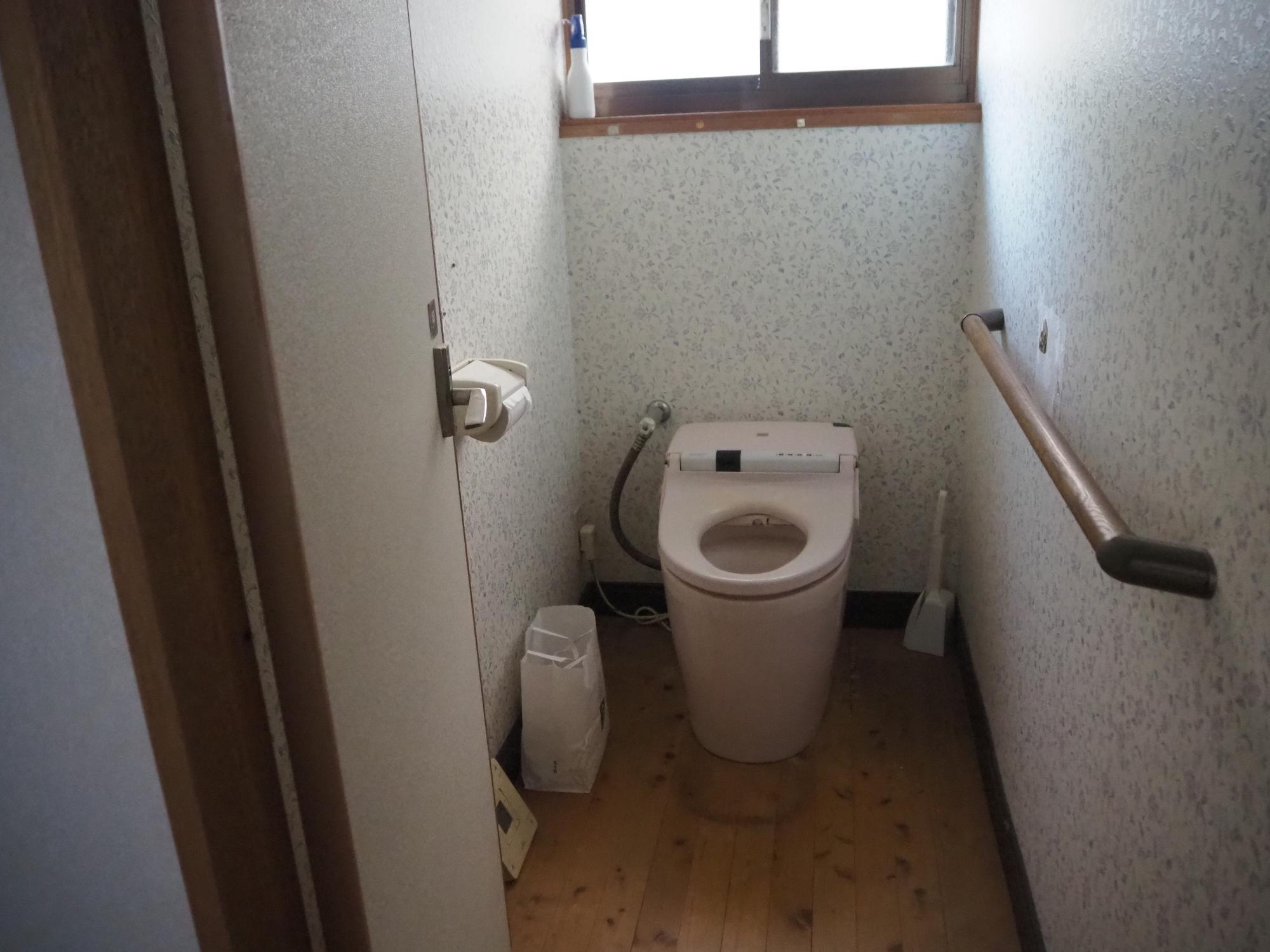 壁には白いクロスが張られ手摺りが取り付けられているピンクの便座のトイレの写真