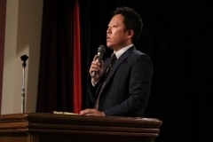 黒いスーツを着た藤田社長が壇上でマイクを持って話をしている写真