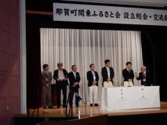 那賀町関東ふるさと会 設立総会・交流会と書かれた幕が張られたステージの上で、スーツを着た男性たちがマイクを持って挨拶をしている写真