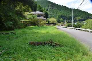 道路沿いの芝生になっている空きスペースの先に民家や山並みが広がっている写真