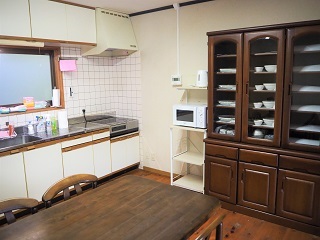 流し台に広めの調理スペースがあり、レンジやポットが置かれた棚、その横には大きな食器棚があり、部屋の中央にテーブルが置かれている台所の写真