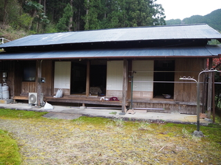 奥に森林がある手前に古い木造窓がある木造平屋の建物の外観写真