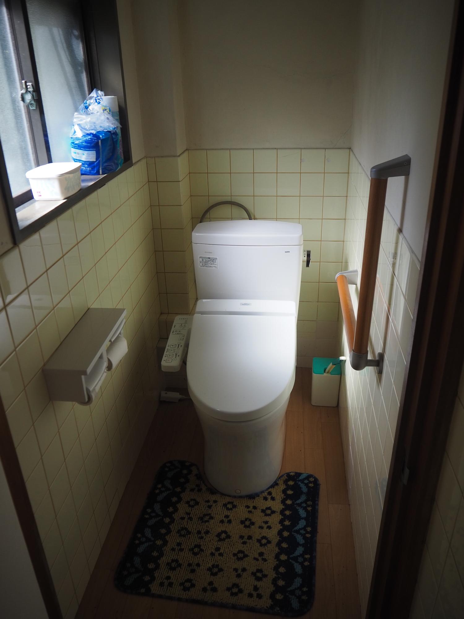 下半分の壁がタイル張りで床がフローリング、右側の壁には手すりが設けられ様式のトイレが設置されているトイレの写真