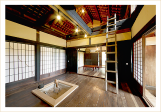 室内で、柱が黒く塗られ、白い障子と板張りの部屋で、天井は梁が見える解放的空間となっており、部屋の真ん中に囲炉裏がある古民家ゲストハウスの写真