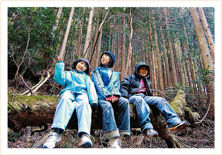 3人の子どもが森の中で倒木に並んで座っている山村留学の風景写真