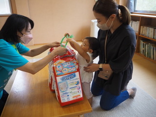 幼稚園のような室内で、おむつのパックを職員の方から受け取っている子どもとお母さんの写真