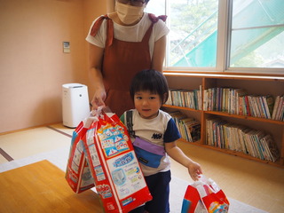 幼稚園のような室内で、おむつのパックを3つ両手に持った子どもとその後ろにいる保護者の写真